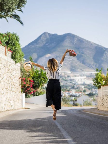 young beautiful woman on vacation jumping. In one hand sandals in the second hand hat.